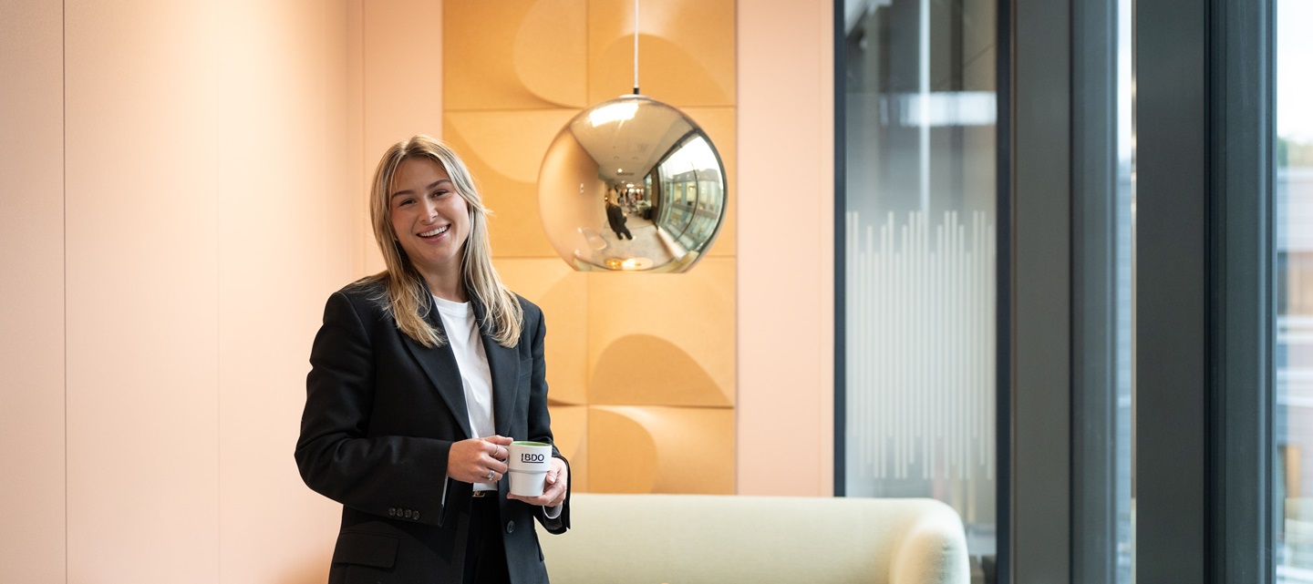 Andrea Munkhaugen står med kaffekoppen i loungeområdet på kontoret hvor hun smiler fra øre til øre